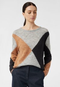 Frau mit dunklen, zurückgebundenen Haaren trägt einen mehrfarbigen Strickpullover mit großen braunen, schwarzen und grauen Farbblöcken sowie dunklen Hosen, die Hände in den Taschen.