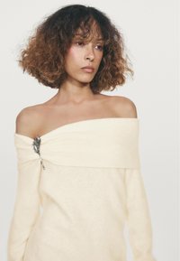 Pull en tricot crème décolleté avec un tissu texturé et un détail de broche argentée sur le côté gauche, présentant un motif de feuille.