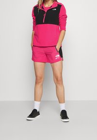 Rosa och svart fleecepullover med halvdragkedja och logotyp, tillsammans med rosa shorts med fickor och logotyp; svarta sneakers och vita strumpor.
