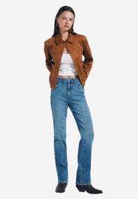 Brun mockaskinnjacka med krage, knappstängning, bärs över en vit topp. Matchad med ljusblå jeans och mörka ankelbooties.