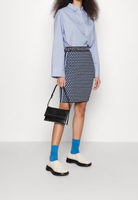 Chemise bleu clair à boutons, jupe crayon noire et blanche à pois, chaussettes bleues, chaussures glissantes crème, et un petit sac à main noir.