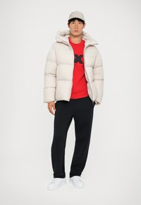 Puffjacka i beige med huva, dragkedja och quiltat mönster. Röd sweatshirt under med svart logga. Mörka byxor och vita sneakers.