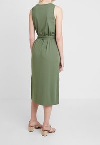 Vestito midi senza maniche verde oliva, con vita a cinta e spacchi laterali. Tessuto morbido, stoffa fluida, silhouette elegante e sandali alla caviglia.