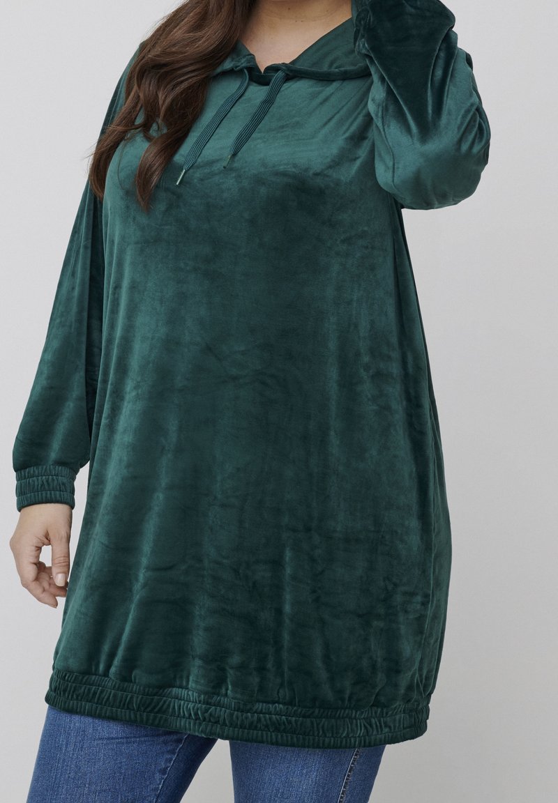 Tunique en velours vert avec capuche, manches longues et poignets élastiques. Elle dispose d'un col avec cordon de serrage et d'une ourlet froncé pour un effet de texture supplémentaire.