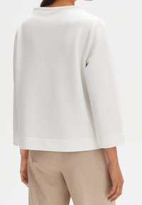 Giacca bianca testurizzata con colletto alto, maniche a tre quarti e orlo arrotondato, abbinata a pantaloni beige su misura.