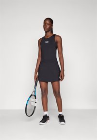 Abito da tennis senza maniche di colore nero con busto aderente e gonna svasata. Presenta il logo EA7, mentre tiene una racchetta da tennis e indossa scarpe sportive.