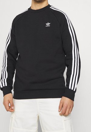 Mand iført en sort Adidas sweatshirt med hvide tre striber på ærmerne og hvide cargobukser mod hvid baggrund.