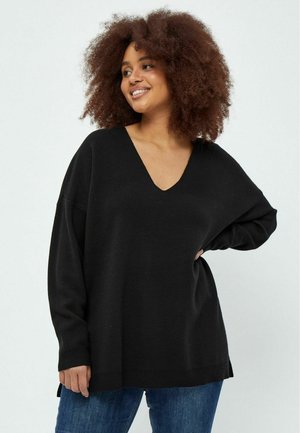 Schwarzer, übergroßer Pullover mit tiefem V-Ausschnitt, langen Ärmeln, gerippten Bündchen und seitlichen Schlitzen. Kombiniert mit blauen Jeans, die einen lässigen Schnitt und Textur zur Schau stellen.
