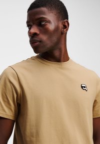 Beige katoenen t-shirt met een klassieke pasvorm, met een klein zwart-wit geborduurd logo op de linkerkant van de borst. Korte mouwen en ronde hals.