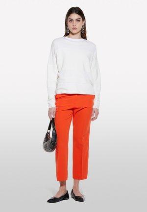 Ein Modell steht vor einem weißen Hintergrund, trägt einen weißen Pullover und leuchtend orangefarbene Hosen und hält eine schwarze Tasche mit silbernen Details.
