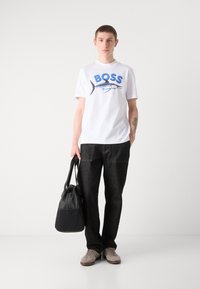 Camiseta blanca de algodón con logotipo "BOSS" en azul y gráfico de marlín, combinada con pantalones de mezclilla negros y zapatillas de ante grises; sostiene una bolsa negra.