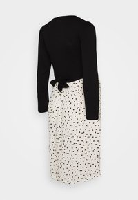 Svart långärmad topp med knytning bak, ihopparad med en krämfärgad kjol med svart prickmönster. Slät tygtextur.