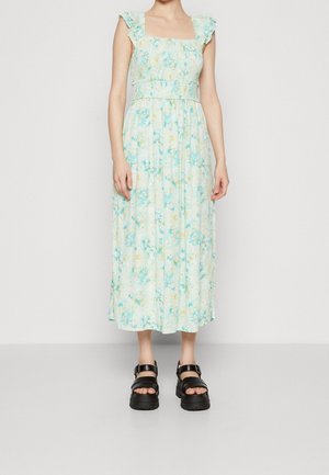 Kvinde iført en ærmeløs, lyseblå midikjole med blomsterprint, flæsekanter på stropperne og sorte platform-sandaler, stående mod en ensfarvet baggrund.