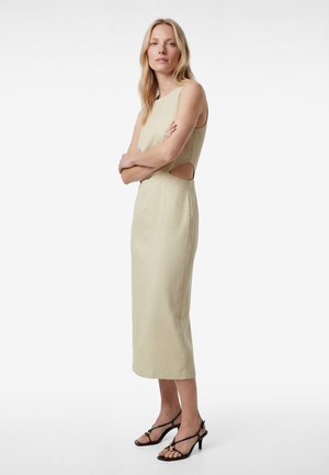 Lyshåret kvinde står med armene over kors iført en ærmeløs beige midi-kjole med sidelommer og sorte hælede sandaler mod en hvid baggrund.