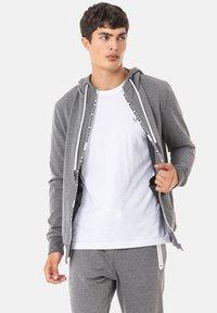 Grau zip-fester Kapuzenpulli mit weißen Kordeln, ausgestattet mit einem kontrastierenden Innenfutter und Logo-Akzenten, getragen über einem weißen T-Shirt und passenden Hosen.