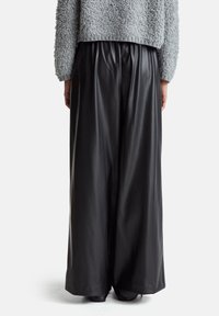 Osoba ubrana w czarne skórzane spodnie o szerokich nogawkach i szary, strukturalny sweter, pokazana od tyłu na jednolitym tle.