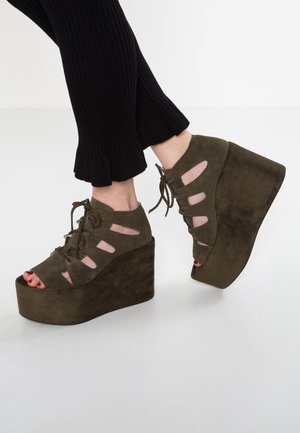 Olivgrüne Wildleder-Plateauabsätze mit offenem Zehenbereich, schnürbarem Vorderteil und Ausschnittdetails. Erhöhte Keilsohle mit strukturiertem Finish.