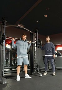 Twee individuen tillen gewichten in een sportschool met grijze en witte sportkleding. Een metalen gewichthefmachine staat op de voorgrond en weerspiegelt de omgevingsverlichting.