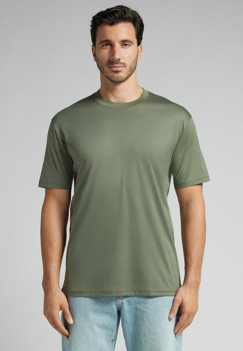 Oliwkowa koszulka z krótkim rękawem, okrągłym dekoltem o gładkiej teksturze i prostym kroju. Połączona z jasnoniebieskimi dżinsami.