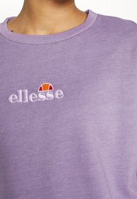 Detailní záběr osoby v levandulovém svetru s bílým a oranžovým vyšitým logem "ellesse" na hrudi.