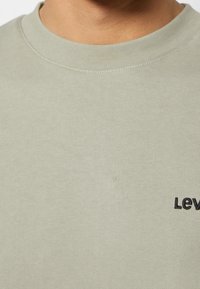 Gros plan sur la partie supérieure de la poitrine et le cou d'une personne portant un t-shirt gris clair à col rond avec un texte noir partiel « Lev » visible.