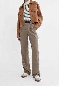 Femme portant une veste cropped marron, un haut rayé blanc et noir, un pantalon beige taille haute, une ceinture noire et des baskets blanches avec des semelles en gomme.