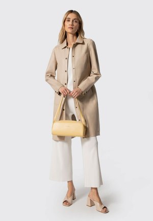 Donna che indossa un cappotto lungo beige, pantaloni bianchi a gamba larga, sandali beige con tacco largo, che tiene una borsa gialla davanti a uno sfondo uniforme.