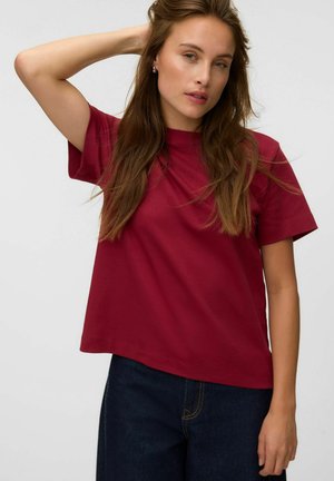 Kvinde med langt brunt hår iført en rød t-shirt og mørke jeans, med den ene arm løftet bag hovedet mod en ensfarvet baggrund.