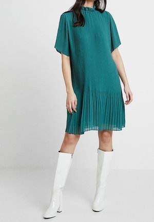 Femme portant une robe plissée bleu sarcelle longueur genou avec manches courtes et bottes blanches montantes jusqu'au genou, debout devant un fond uni.
