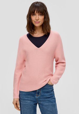 Roze gebreide trui met een V-hals, lange mouwen en een relaxte pasvorm. Onder de trui is een donkerblauw T-shirt met ronde hals zichtbaar. Draagt met blauwe spijkerbroek.