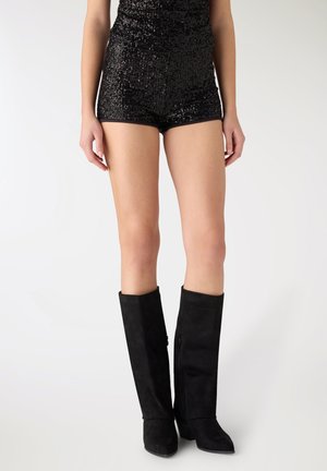 Donna che indossa pantaloncini neri con paillettes e stivali di camoscio neri al ginocchio, in piedi contro uno sfondo chiaro e uniforme.