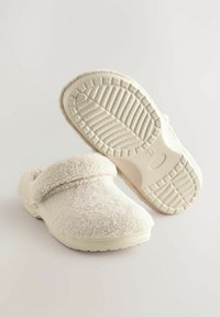 Zapatillas blancas y suaves con una textura blanda, punta redonda y una correa acolchada en la parte superior; suelas de goma con un diseño de agarre.
