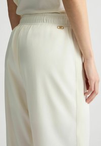 Crème-kleurige, lichte broek met een elastische tailleband en zijzakken; heeft een klein logo in goudkleurige toon aan de achterkant.