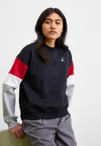 Sudadera negra con un logo de Jordan en blanco, mangas con acentos rojos y blancos, y puños elásticos grises. El material parece suave y texturizado.