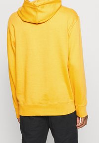 Persona con un sudadero con capucha amarillo brillante, visto desde atrás, combinado con pantalones negros sobre un fondo neutro.