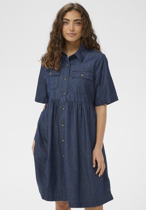 Göndör hajú nő rövid ujjú, gombos, sötét farmer anyagból készült, mellzsebes ruhában, egyszínű világos háttér előtt állva.