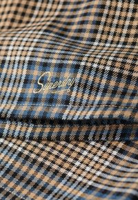 Tissu à carreaux dans des tons bleu, noir et beige avec une texture douce. Comprend un logo brodé en or "Superdry" près de la couture.