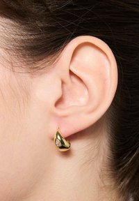 Złote kolczyki w gładkim, kroplowym kształcie z małym przezroczystym akcentem kamienia szlachetnego. Umieszczone na małżowinie usznej, prezentujące wypolerowane wykończenie.