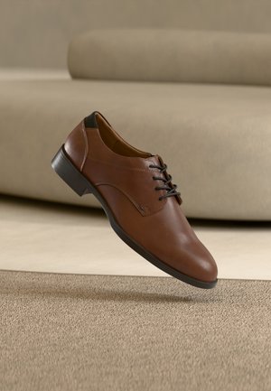 Chaussure habillée pour homme en cuir marron avec lacets noirs et semelle noire, flottant au-dessus d'un tapis beige texturé et d'un canapé beige en arrière-plan.