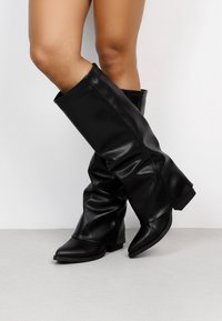 Botas negras de imitación de cuero hasta la rodilla con un diseño ligeramente holgado, punta afilada y tacón cuadrado. Textura suave con detalles de costura lateral.