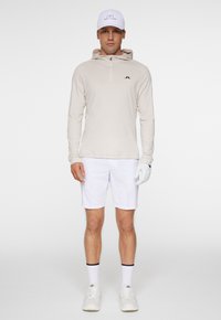 Beige långärmad tröja med huva och halvdragkedja, ihop med vita shorts. Vit keps med en logotyp, och handskarna är ljusgrå. Vita sneakers.