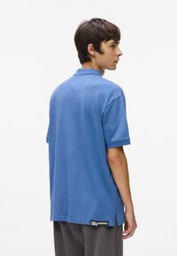 Μπλε πόλο μπλουζάκι με κοντά μανίκια, επίπεδο γιακά και πλαϊνά σκισίματα. Φέρει μια μαύρη ετικέτα λογότυπου στην αριστερή πλευρά κοντά στο στρίφωμα.