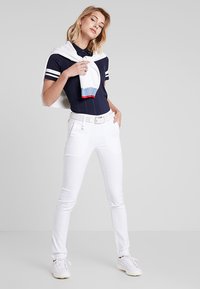 Marineblauwe polo met witte strepen, gecombineerd met witte slim-fit broeken en een witte sweater om de schouders geknoopt. Witte sneakers maken de look af.