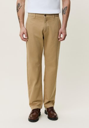 Mann iført beige bukser med rette ben, brune lær sko og hvit skjorte, står med hendene avslappet langs siden.
