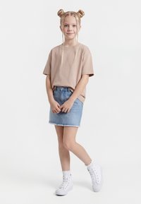 Jauna meitene stāv, sakrustotām rokām, ģērbusies b ēša krāsas T-kreklā, džinsa svārkos, baltās zeķēs un baltās augstās teniskurpēs, ar pinušām matu bulciņām.