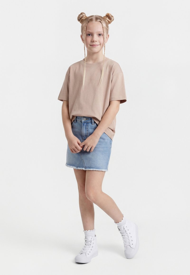 Jauna meitene stāv, sakrustotām rokām, ģērbusies bēša krāsas T-kreklā, džinsa svārkos, baltās zeķēs un baltās augstās teniskurpēs, ar pinušām matu bulciņām.