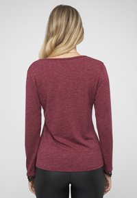 Bordeauxrotes Langarmshirt mit rundem Ausschnitt und lockerem Sitz, gefertigt aus einem weichen, strukturierten Stoff; kombiniert mit schwarzen, eng anliegenden Hosen.