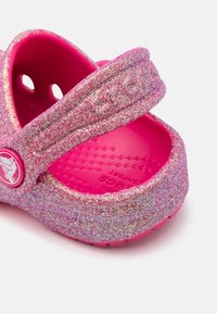 Zoccolo rosa glitterato con una cintura larga, superficie testurizzata e un logo ovale. Presenta fori di ventilazione e un design della soletta imbottita.