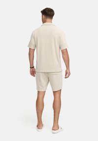 Helles beige Kurzarmhemd und passende Shorts aus strukturiertem Stoff. Die Rückseite zeigt einen Kragen und einen lockeren Schnitt, mit einer einzelnen Gesäßtasche.