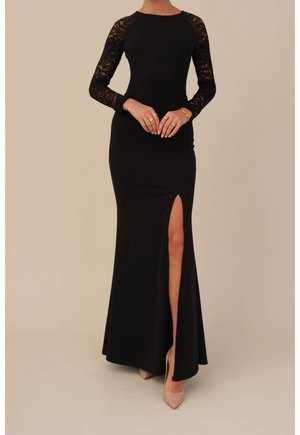 Zwarte jurk tot op de vloer met kanten lange mouwen, hoge split tot aan de dij, beige hakken en gouden ringen aan de vingers tegen een neutrale achtergrond.